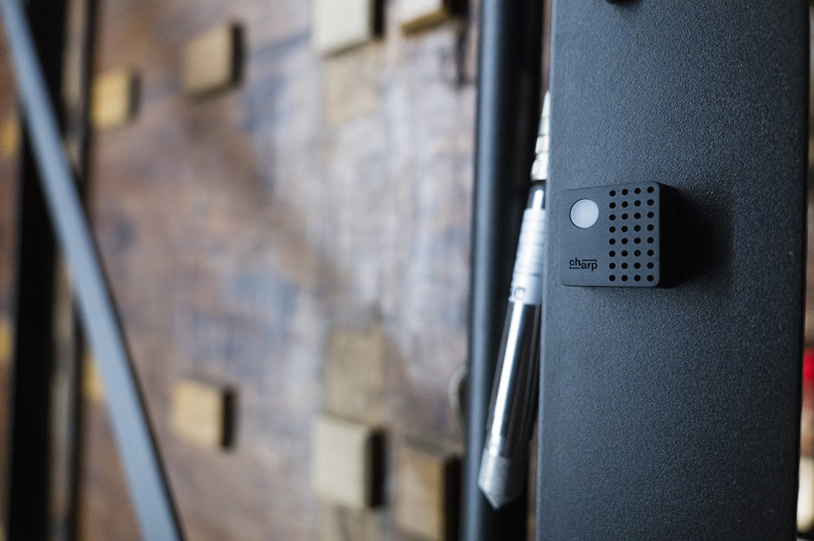 A black Charp Mini logger fixed to the dark frame of a display case in front of a brick wall.