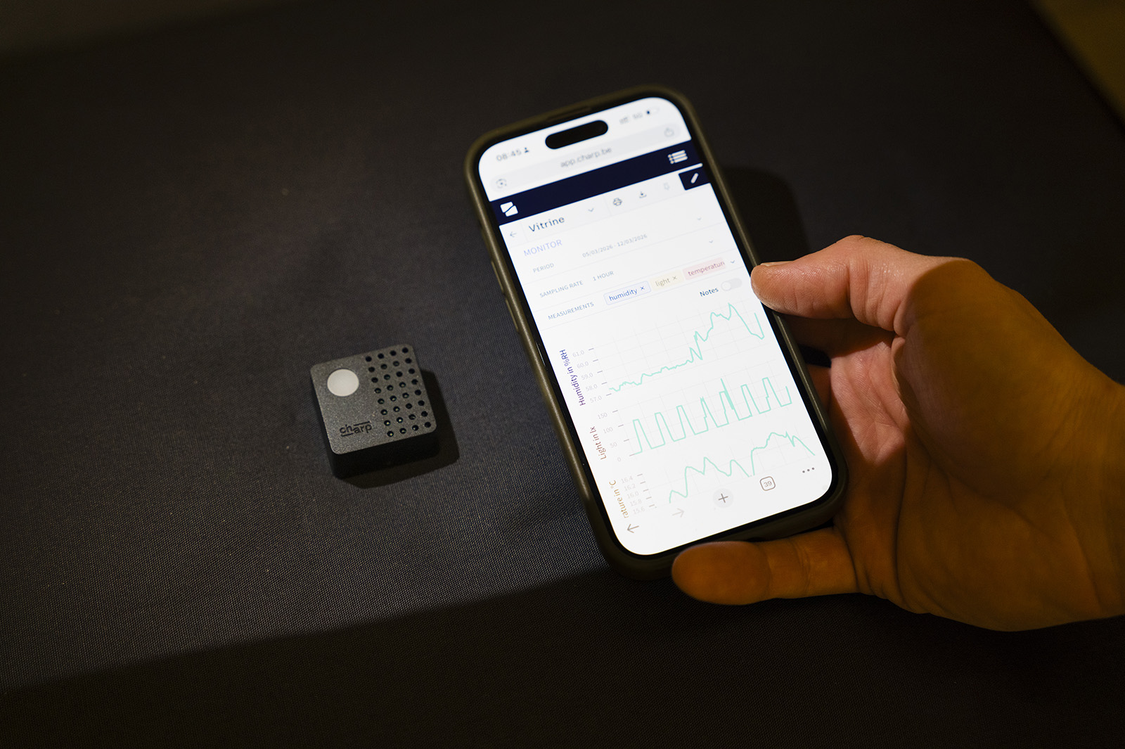 A hand holding a phone showing Charp temperature and humidity graphs, with a Maxi logger on the surface alongside.